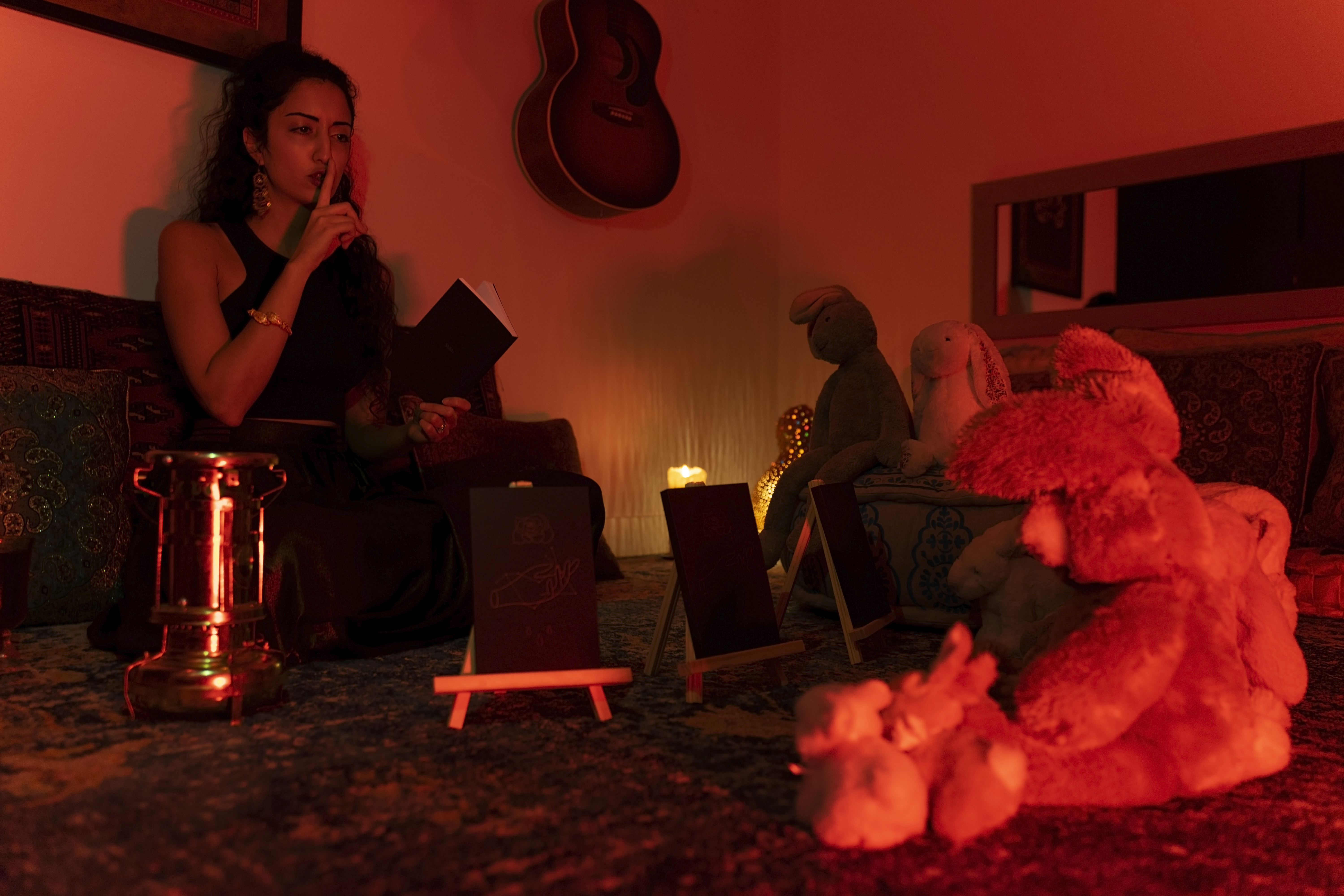 a photo of Samaneh sitting on the floor of a dark room decorated by a rug and pillows and shushing a bunch of stuffed animal rabbits so she can read them her book.